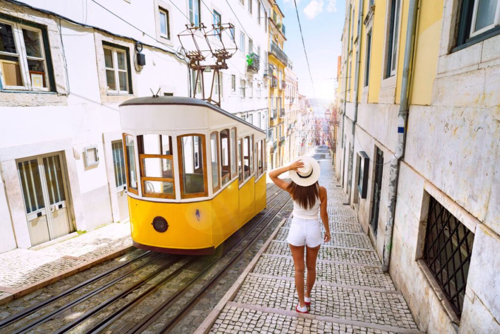 Mulher turista andando em ruas estreitas da cidade velha de Lisboa, ao lado do bonde funicular amarelo retrô em um dia ensolarado no verão europeu.