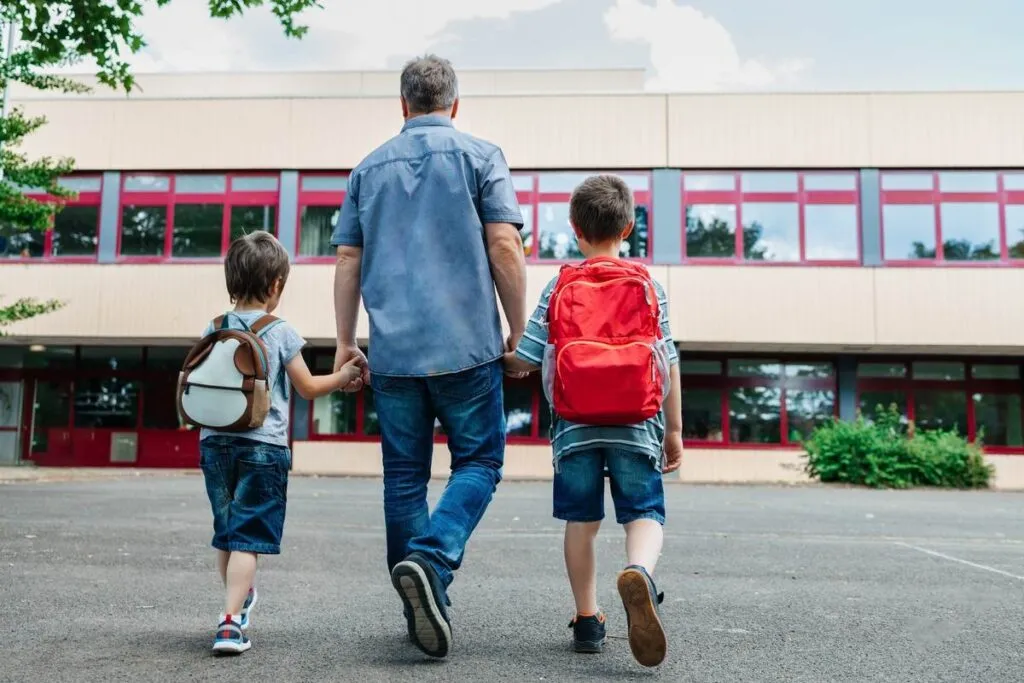 Duas crianças com mochilas nas costas segurando as mãos do pai e indo para escola.