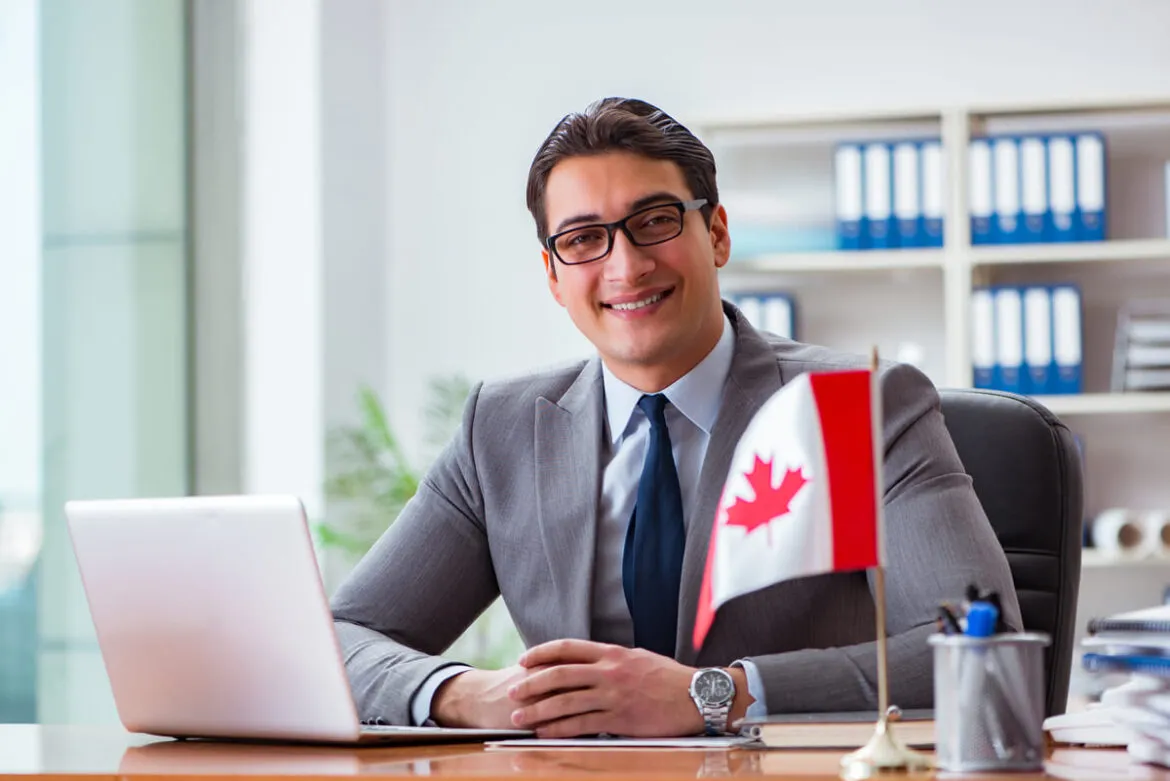 Um homem branco sentado diante de uma mesa. Na imagem há uma bandeira canadense que simboliza como trabalhar no Canadá.