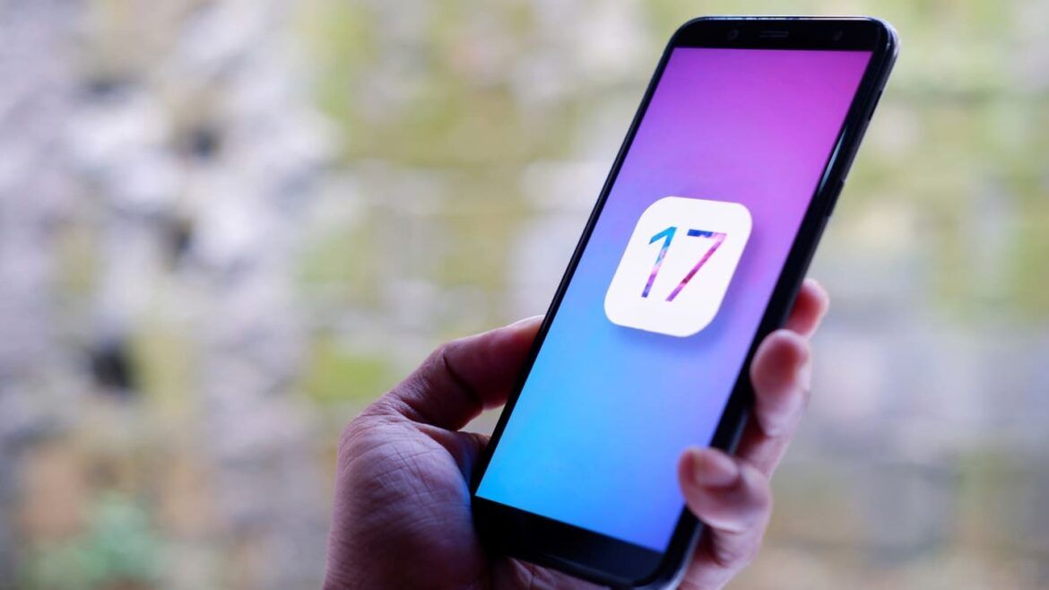 Imagem de uma mão branca segurando um iPhone com o texto "iOS 17" na tela.