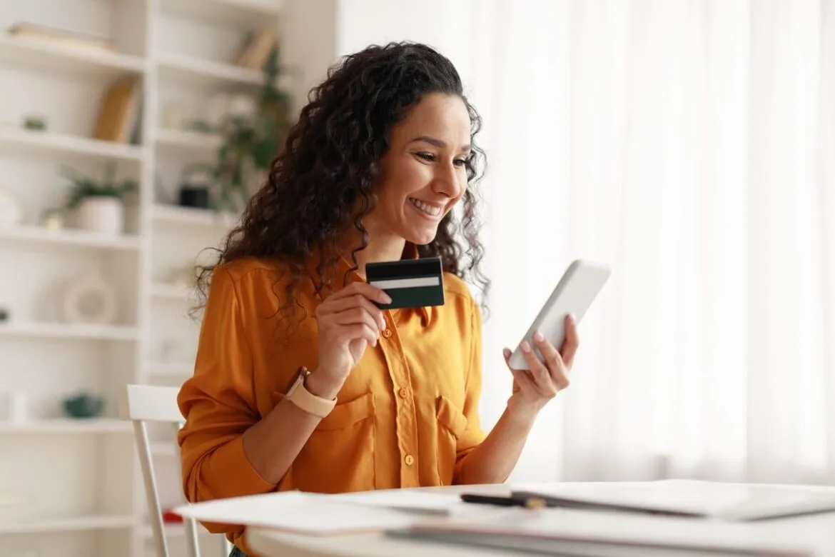 Mulher segurando o cartão Estrela internacional conta corrente e acessando a conta online no smartphone.