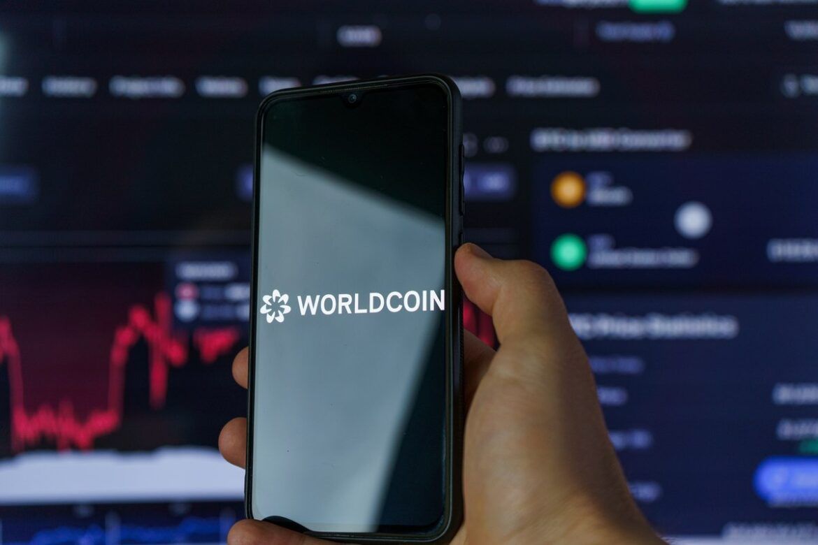Mão de homem segurando smartphone com o logo do WorldCoin em uma tela preta.