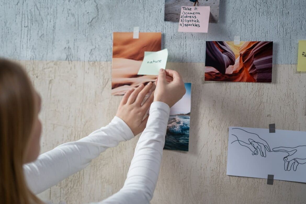 Mãos de uma mulher prendendo um post it em uma parede, juntamente a algumas imagens e outros post its, para criar um moodboard.