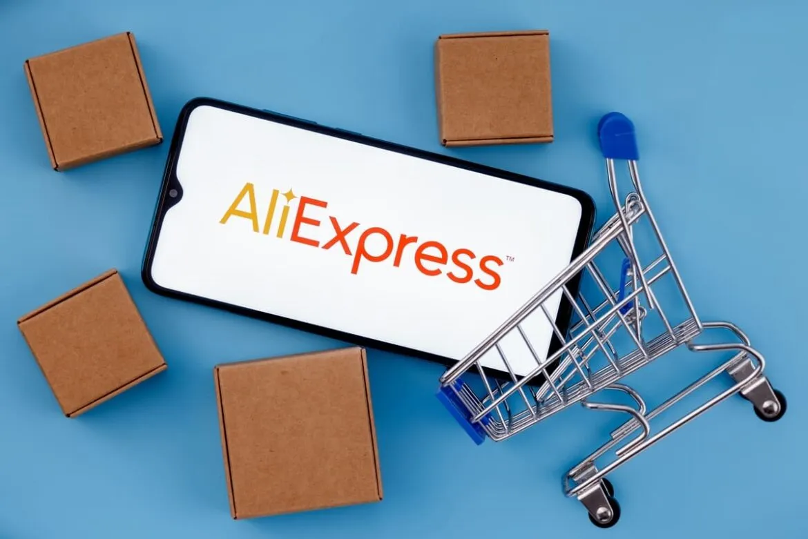 Telefone escrito "AliExpress" em um fundo azul, com pequenas caixas de papelão e um carrinho de compras simbolizando aquisições de produtos internacionais feitos no e-commerce, que agora aderiu ao Programa Remessa Conforme.