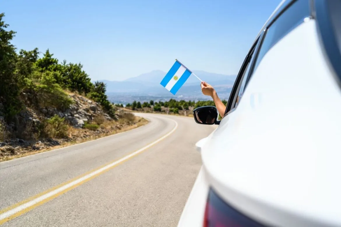 Mulher dirigindo carro branco e segurando bandeira da Argentina.