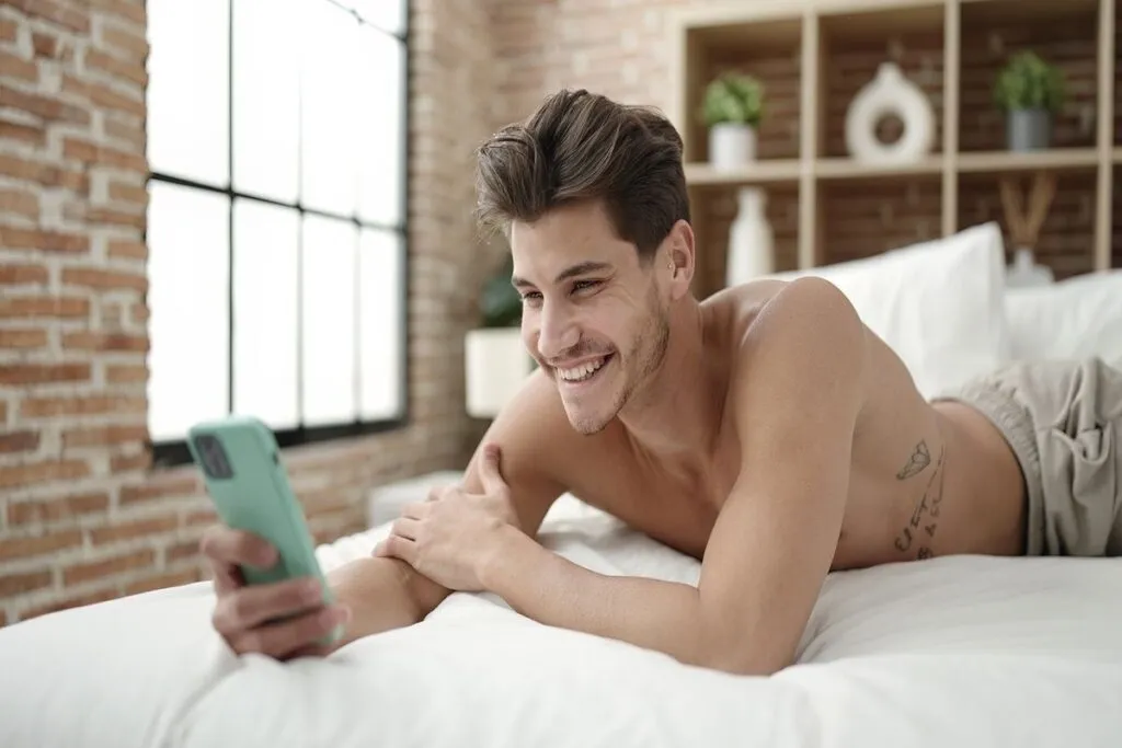 Homem sem camisa sorrindo ao olhar a tela do celular. 