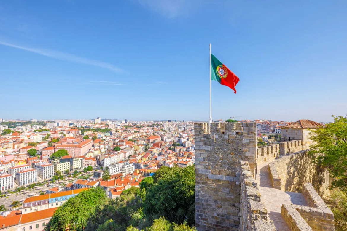 Imagem de um castelo em Portugal com a bandeira portuguesa com uma cidade ao fundo. Portugal está passando por uma alta de 505% de denúncias de xenofobia nos últimos anos.