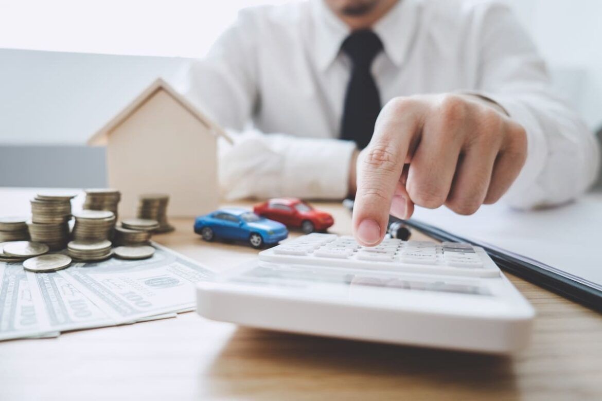 Imagem de um homem fazendo cálculos na calculadora, com moedas, carrinhos de brinquedo e uma pequena maquete de uma casa para ilustrar um texto sobre alienação fiduciária.