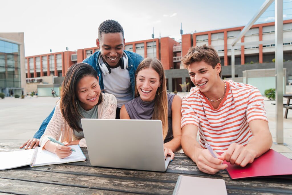 Estudantes internacionais reunidos ao redor de uma mesa, olhando diretamente para um notebook. Todos aproveitaram programas de intercâmbio gratuito.