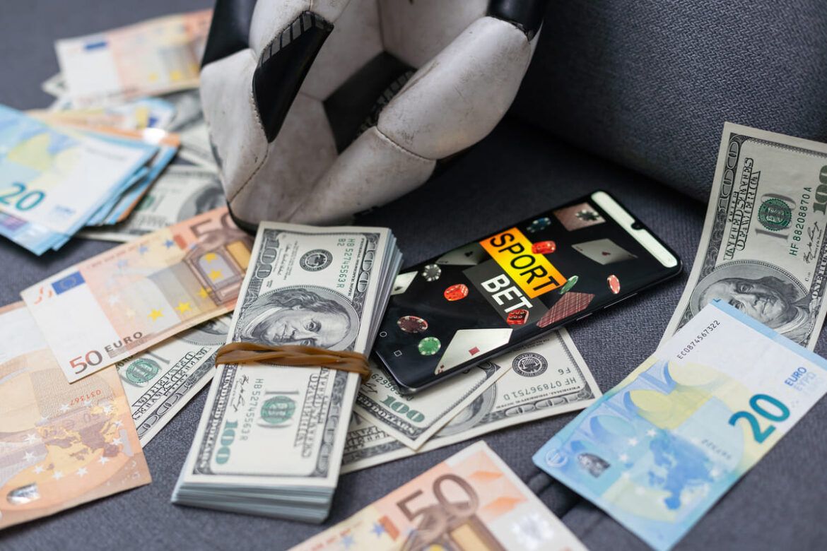 Notas de dinheiro, uma bola de futebol murcha e um celular com a loga marca de uma casa de apostas, para ilustrar um texto sobre a taxa aposta esportiva