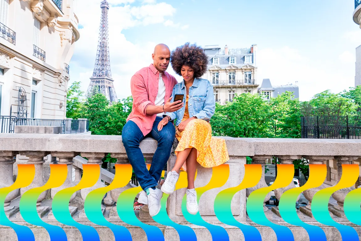 Um casal sentado em uma mureta, em um belo dia de sol, segurando o celular e com a Torre Eiffel ao fundo