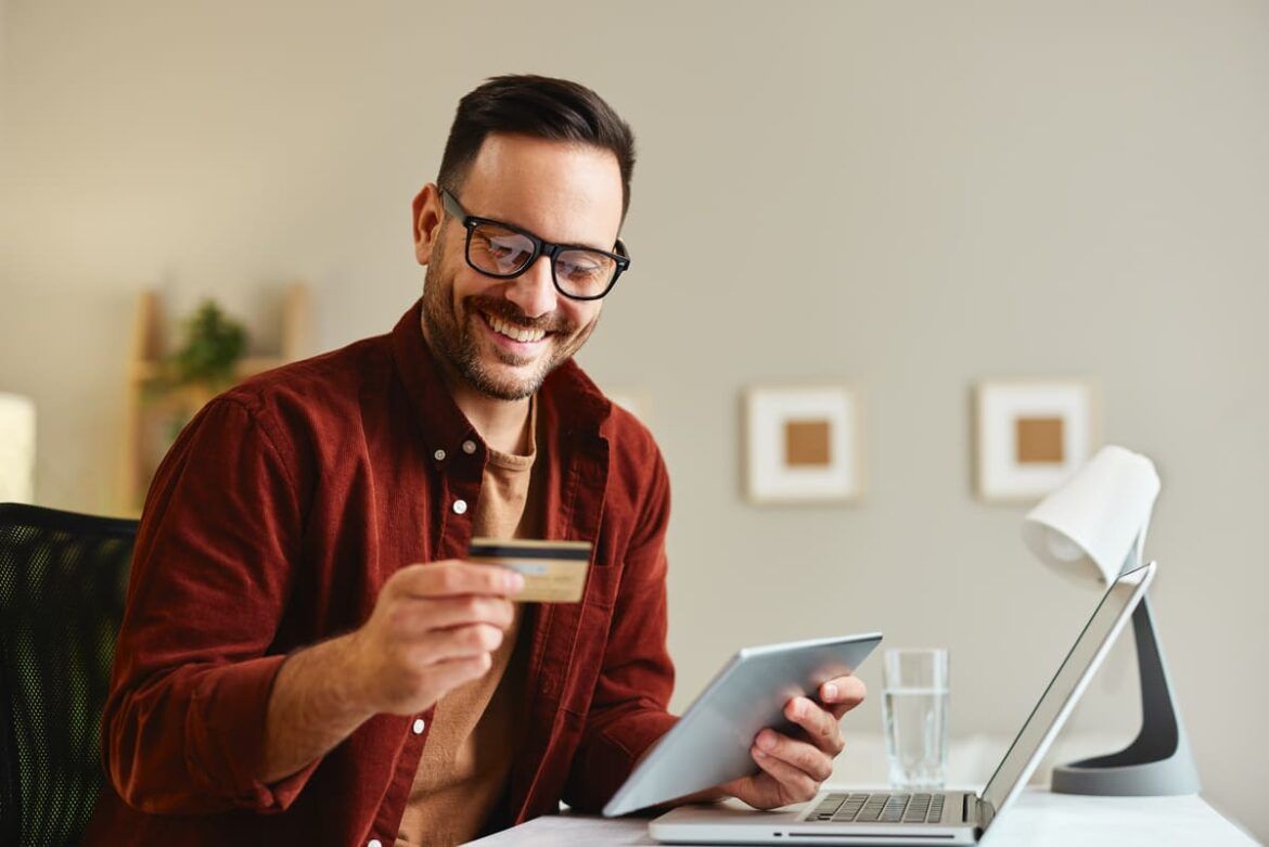 Homem olhando para cartão de crédito em frente ao seu computador e tablet para ilustrar um texto sobre juros rotativo do cartão de crédito