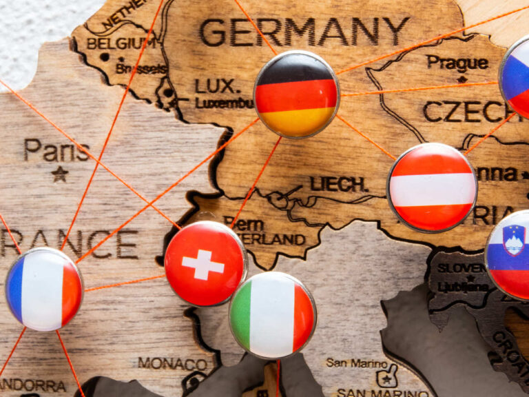 A imagem mostra um mapa da Europa com diversas bandeiras de países, representadas por botões coloridos, marcando os locais no mapa. As bandeiras estão conectadas por linhas vermelhas, sugerindo alguma forma de rede ou conexão entre os países, que pode ser associado ao Espaço Schengen