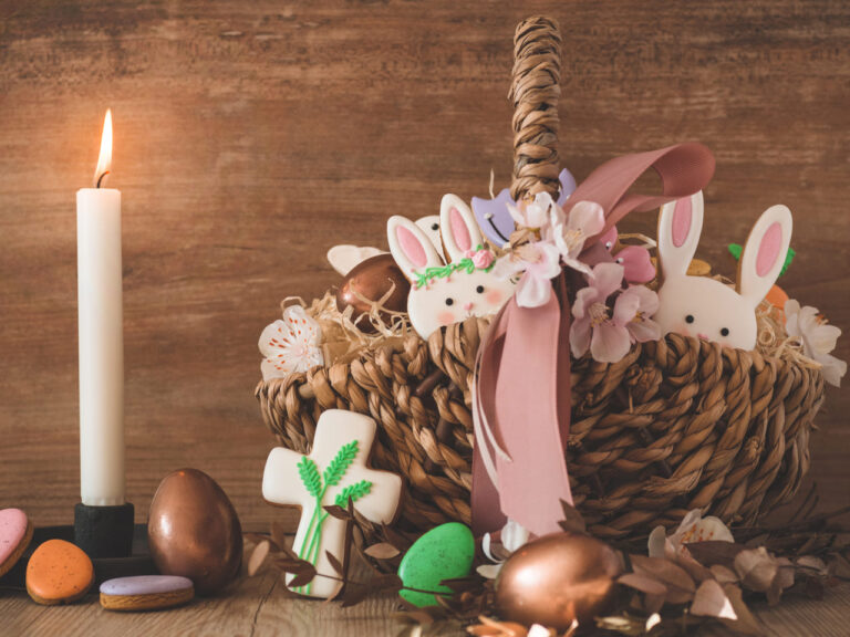 Cesta de Páscoa feita de palha, decorada com flores e um laço rosa. Dentro da cesta, há figuras de coelhos de açúcar, ovos de chocolate e uma cruz decorada com flores e folhas. Ao lado da cesta, há uma vela acesa e alguns doces coloridos, como ovos e biscoitos, espalhados sobre a superfície de madeira.
