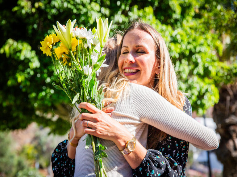 Mãe muito feliz abraçando sua filha ao receber flores amarelas e uma mensagem para o Dia das Mães