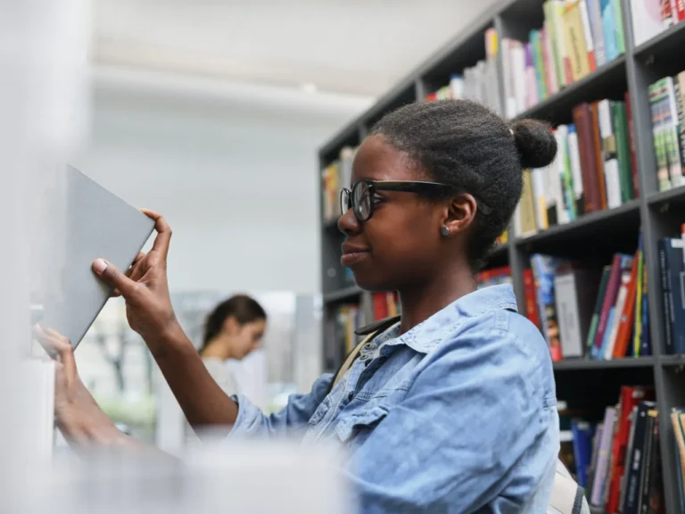 Jovem negra de óculos retira um livro da estante em uma biblioteca, representando o acesso à educação e a iniciativa MEC Livros.