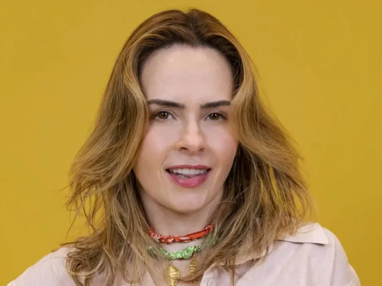 Retrato frontal de Ana Paula Renault sorrindo contra um fundo amarelo vibrante, destacando seu visual com colares coloridos.